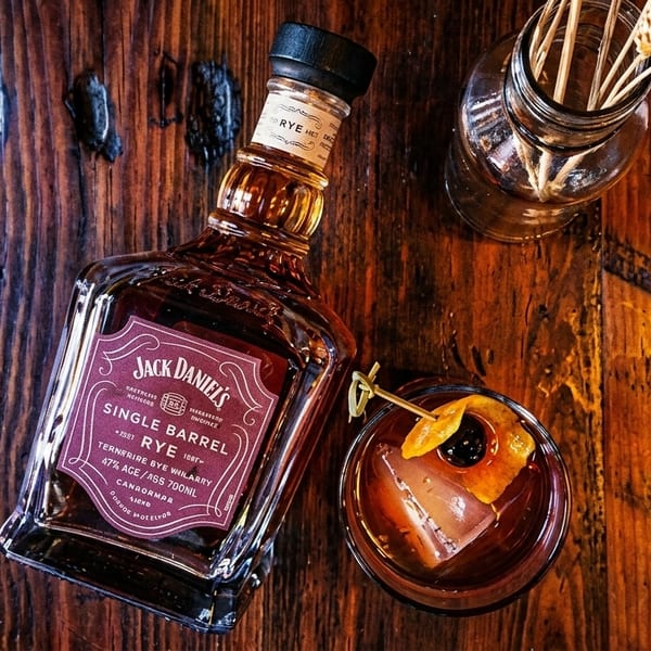 Jack Daniel's Single Barrel Rye bottle next to a cocktail with an orange peel on a wooden table.