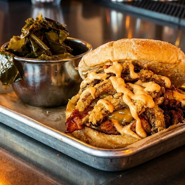 A fried chicken sandwich with sauce and a side of collard greens on a metal tray.