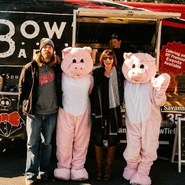 Two people and two pig mascots stand in front of the BowTie Barbecue food truck.