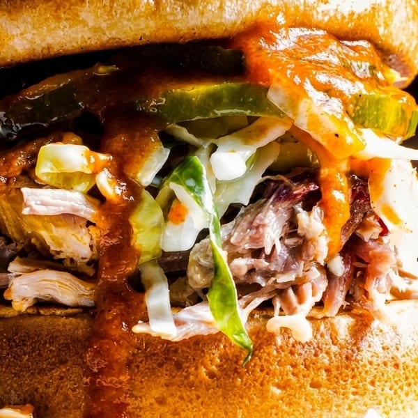 Close-up of a pulled pork sandwich with coleslaw, pickles, and barbecue sauce on a brioche bun.